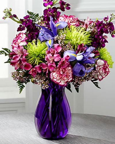 Blooming with rich color and undeniable texture, this stunning bouquet expresses love and gratitude with each perfect flower. Blue Iris, fragrant purple gilly flower, purple Peruvian Lilies, purple bi-color carnations, green spider chrysanthemums, clusters of pink wax flower, and lush greens create a fascinating display presented in a deep purple glass vase with modern lines to create a sophisticated look.
