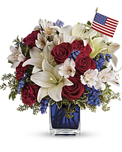 A patriotic pick to send a brave veteran, decorate your Fourth of July picnic, or celebrate Memorial Day. Lush red, white and blue flowers are presented in a deep blue glass cube vase along with an American flag.
Red roses, white asiatic lilies, white alstroemeria, blue statice and red carnations are gathered with green pitta negra, seeded eucalyptus and a small American flag in a cobalt blue glass cube.