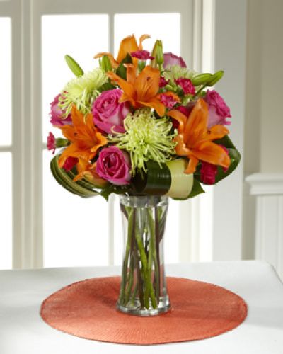 Hot pink bi-colored roses, orange Asiatic Lilies, green Fuji Chrysanthemums, hot pink mini carnations, tropical leaves, and lush greens are brought together to impress, presented in a clear glass vase. Bright, beautiful, and ready to get the party started, this bouquet creates a unique surprise to absolutely delight your recipient with each sunlit bloom.