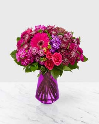 Our Blushing Bouquet is sure to turn heads with its explosion of vibrant purple and hot pink tones. There’s no comparison to the stunning array of gerberas, mini carnations, spray roses, button poms, and stock, complemented by touches of salal, placed in a vibrant purple glass vase.